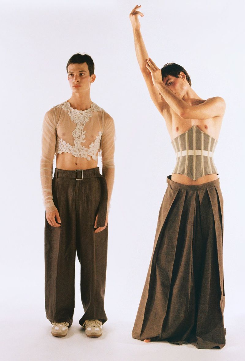 Two men in avant-garde fashion, one shirtless with a corset, the other in a sheer top and wide-leg pants, posing against a white backdrop.