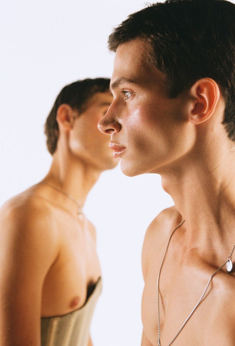 Studio portrait of two young adult men in a bright photo studio. One man in a corset. They are wearing necklaces and have neutral expressions.