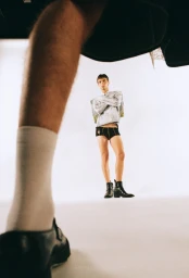 Low angle fashion photograph of a male model wearing shorts and boots, framed past another person's leg in the foreground.