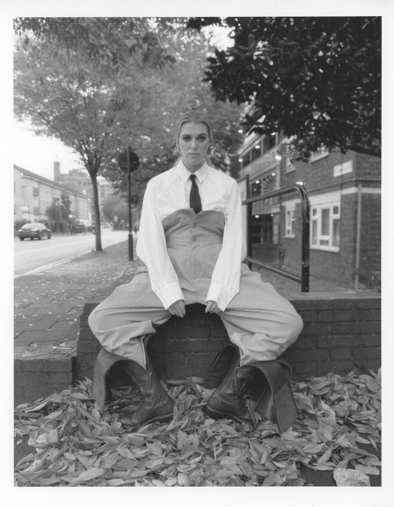 Black and white portrait of a woman in overalls sitting on a stone structure covered in leaves in an outdoor urban environment.