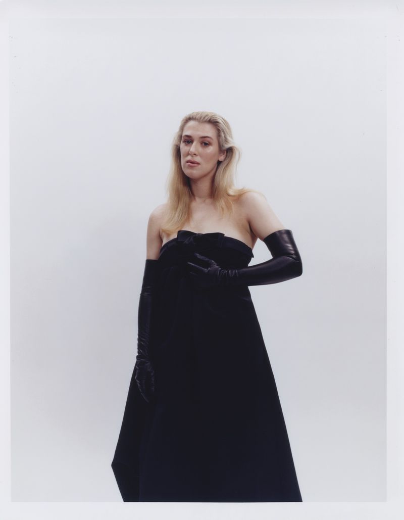 Elegant adult woman poses in a strapless black dress and opera gloves against a stark white backdrop in a fashion-style studio portrait.