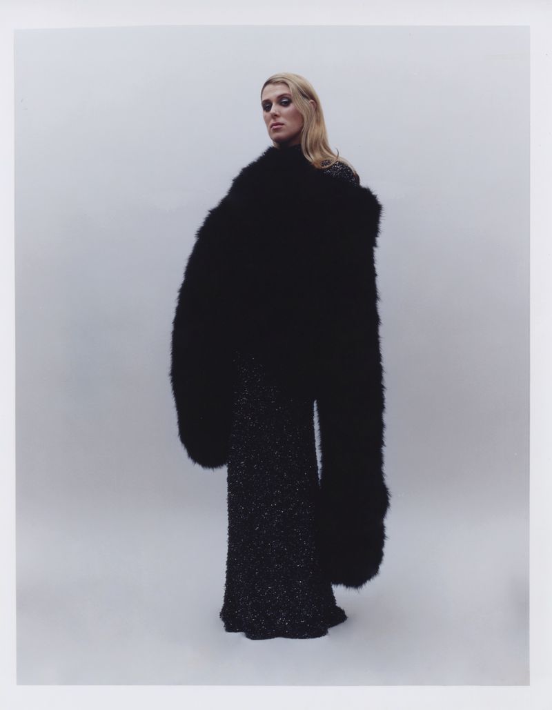 Fashion shot of a blonde adult female model in a flowing black textured dress and oversize fur coat against a light grey backdrop.