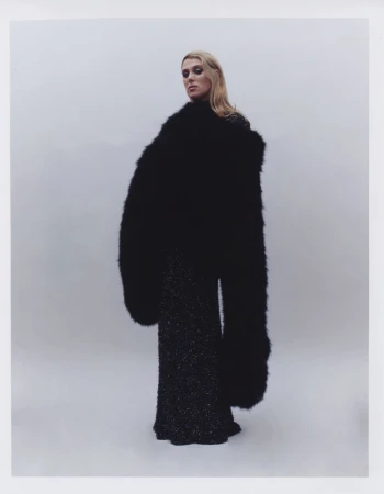Fashion shot of a blonde adult female model in a flowing black textured dress and oversize fur coat against a light grey backdrop.
