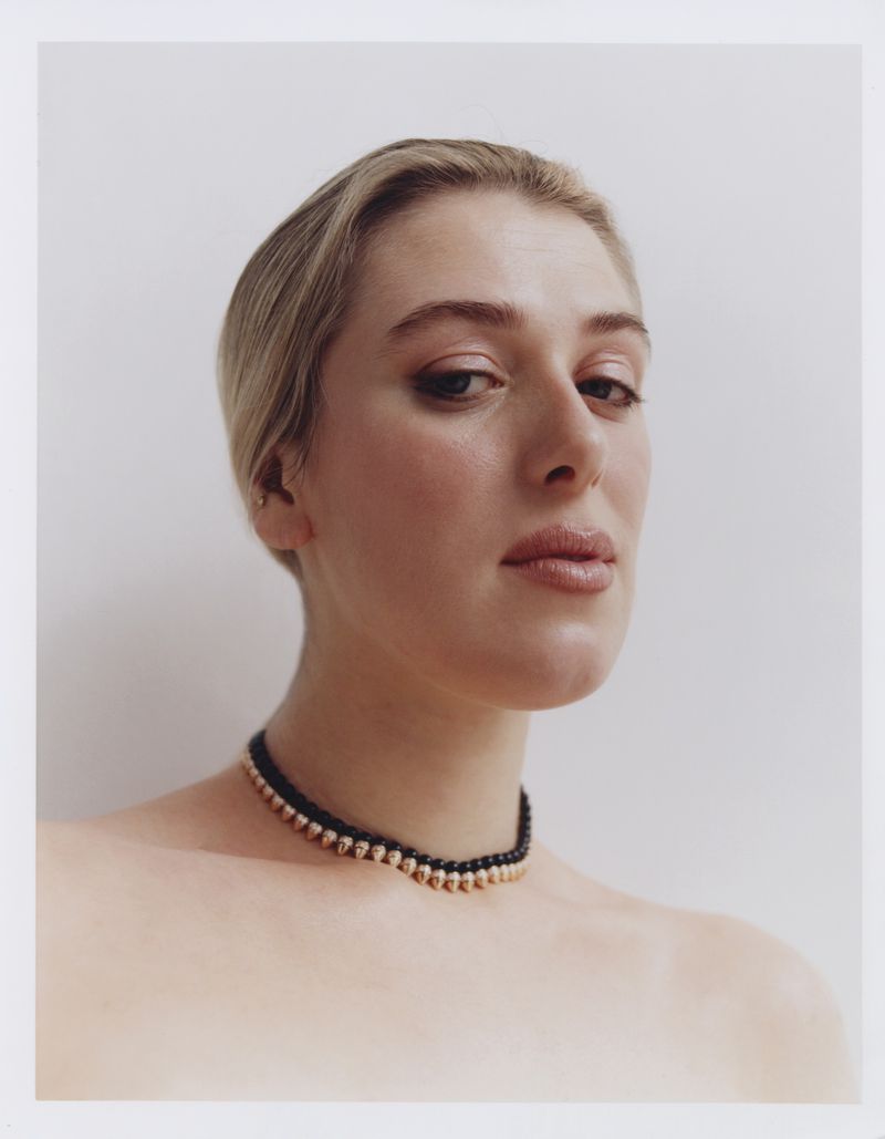 Close-up portrait of a young woman with fair skin and blonde hair, wearing a black and gold beaded choker against a white background.