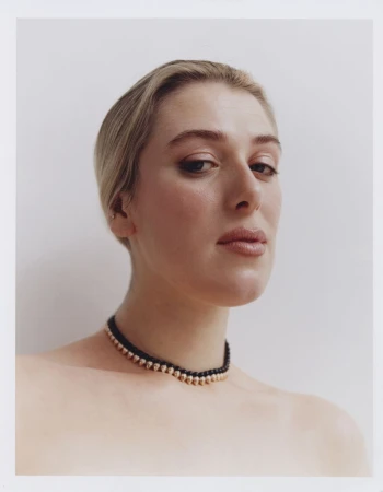 Close-up portrait of a young woman with fair skin and blonde hair, wearing a black and gold beaded choker against a white background.
