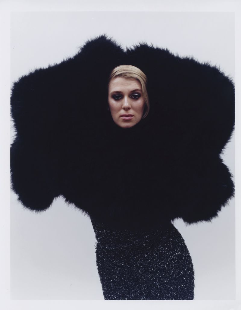 Portrait of a woman in a black, sparkling dress and oversized black fur collar against a neutral background in a studio.