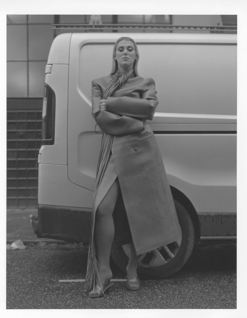 Black and white fashion photograph of an adult woman in a formal dress posing in front of a white van in an urban environment.