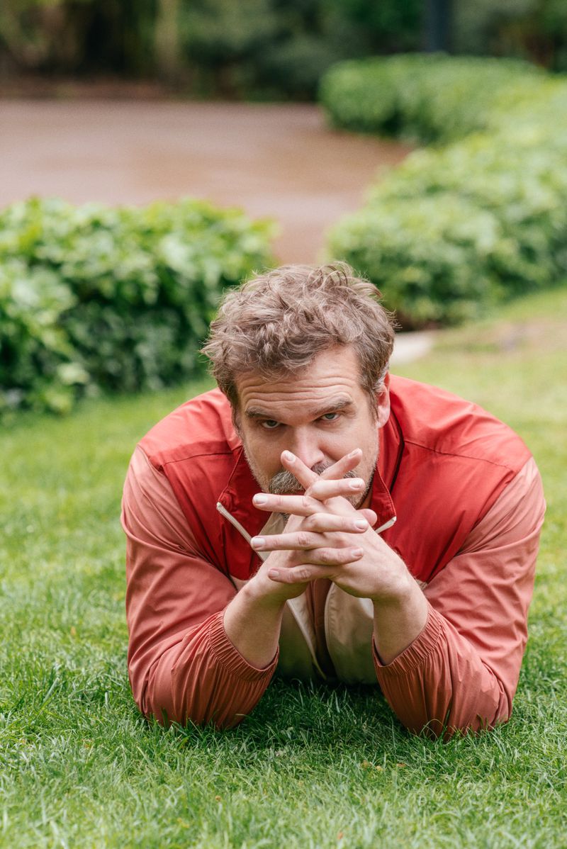 David Harbour poses on grass in a red jacket, looking directly at the camera with his hands clasped together; green plants in background.