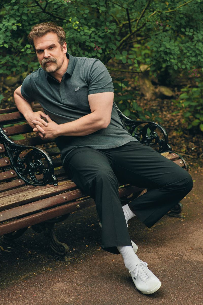 David Harbour poses in a park on a bench. The Stranger Things actor wears a polo shirt and pants while seated with crossed arms.