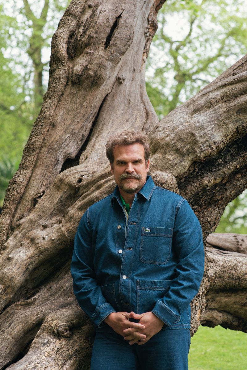 David Harbour poses near a large tree outdoors. He wears a blue denim jacket and jeans, with green peeking from beneath the collar.