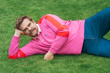 David Harbour wearing a pink polo shirt with red stripes and blue jeans reclining on a green grass lawn
