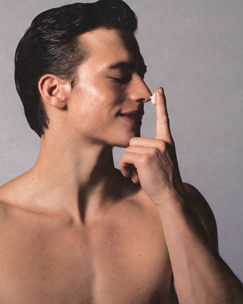 Attractive bare-chested man with a dab of white cream on his nose smiles against a neutral background in soft, even studio lighting.