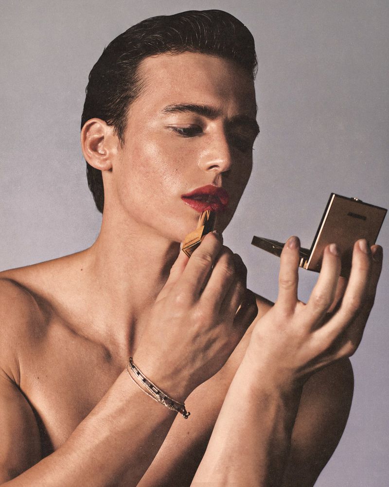 Close-up of a young adult male applying red lipstick while holding a mirror, set against a grey background in a beauty editorial style.