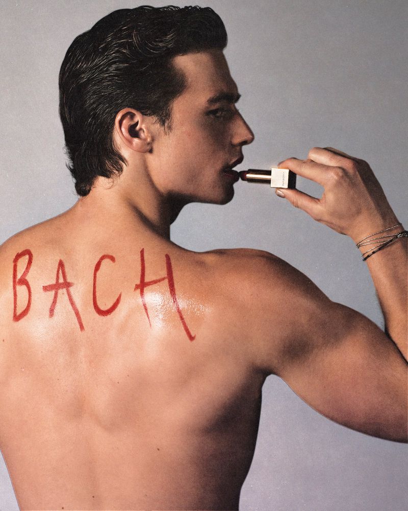 Nude man with back to camera holds lipstick tube to his lips; painted red lettering 'BACH' across his back; grey background.