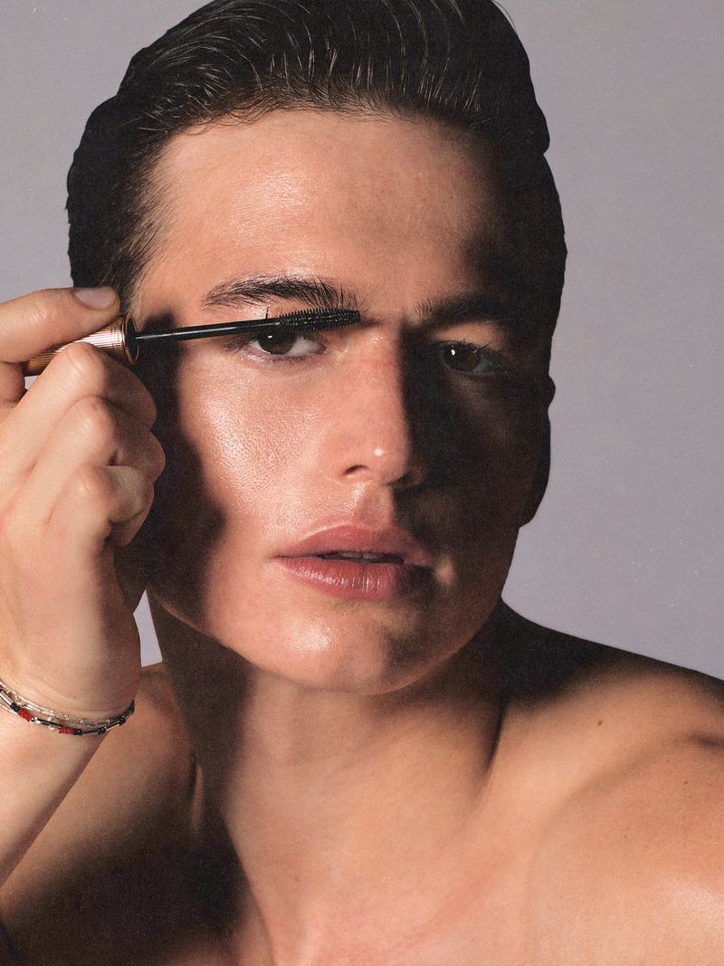 Close-up of a light-skinned adult male applying mascara to his eyelashes in a beauty-style portrait against a grey gradient background.