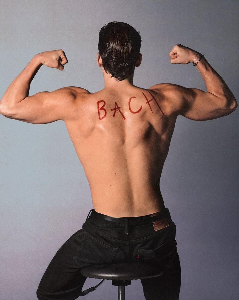 Shirtless man with muscular build poses with back to camera, flexing arm muscles, sitting on stool in front of a grey background.