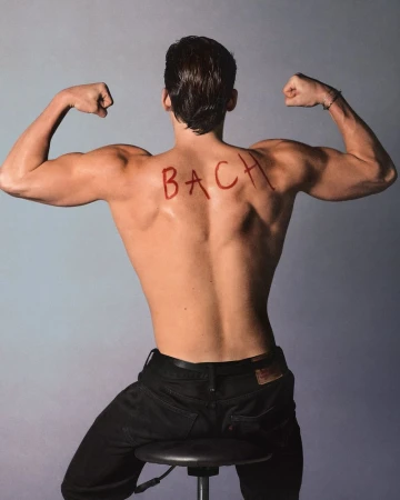 Shirtless man with muscular build poses with back to camera, flexing arm muscles, sitting on stool in front of a grey background.