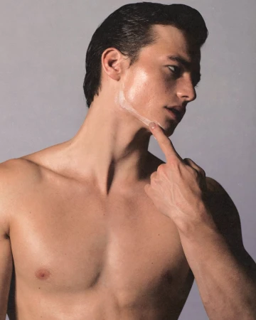 Close-up portrait of a young adult male with skin treatment on his neck, pointing to his face, against a neutral gray background.