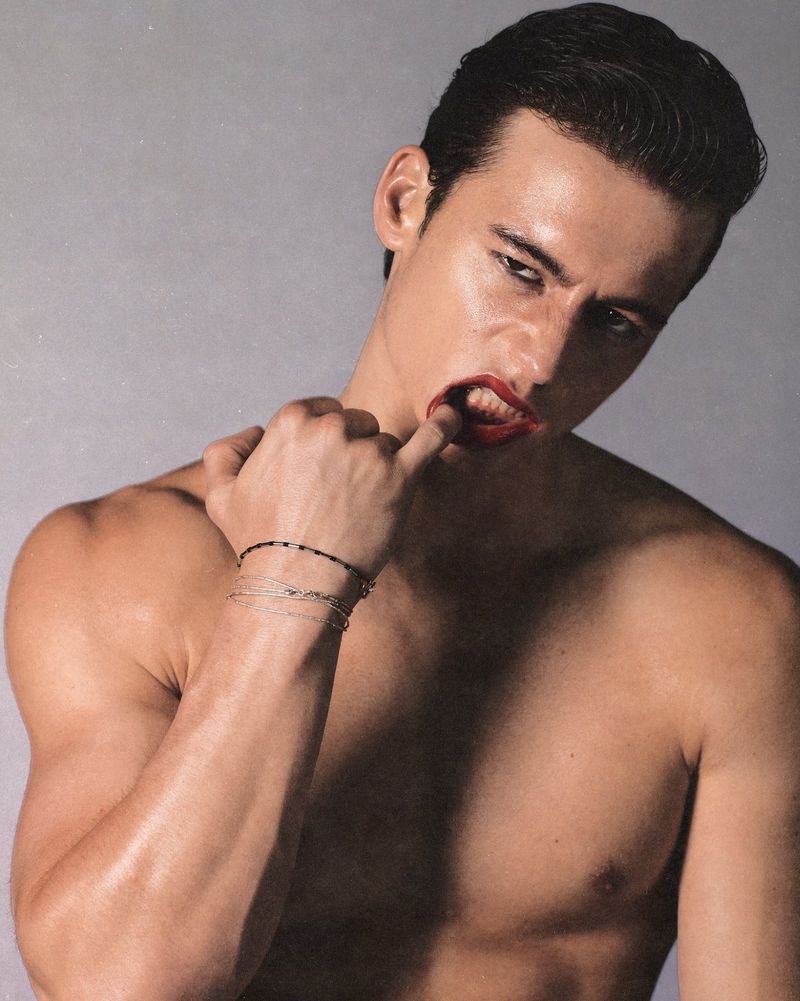 Artistic studio shot of a shirtless man with red lipstick biting his finger, posing against a neutral backdrop. Features bracelets.