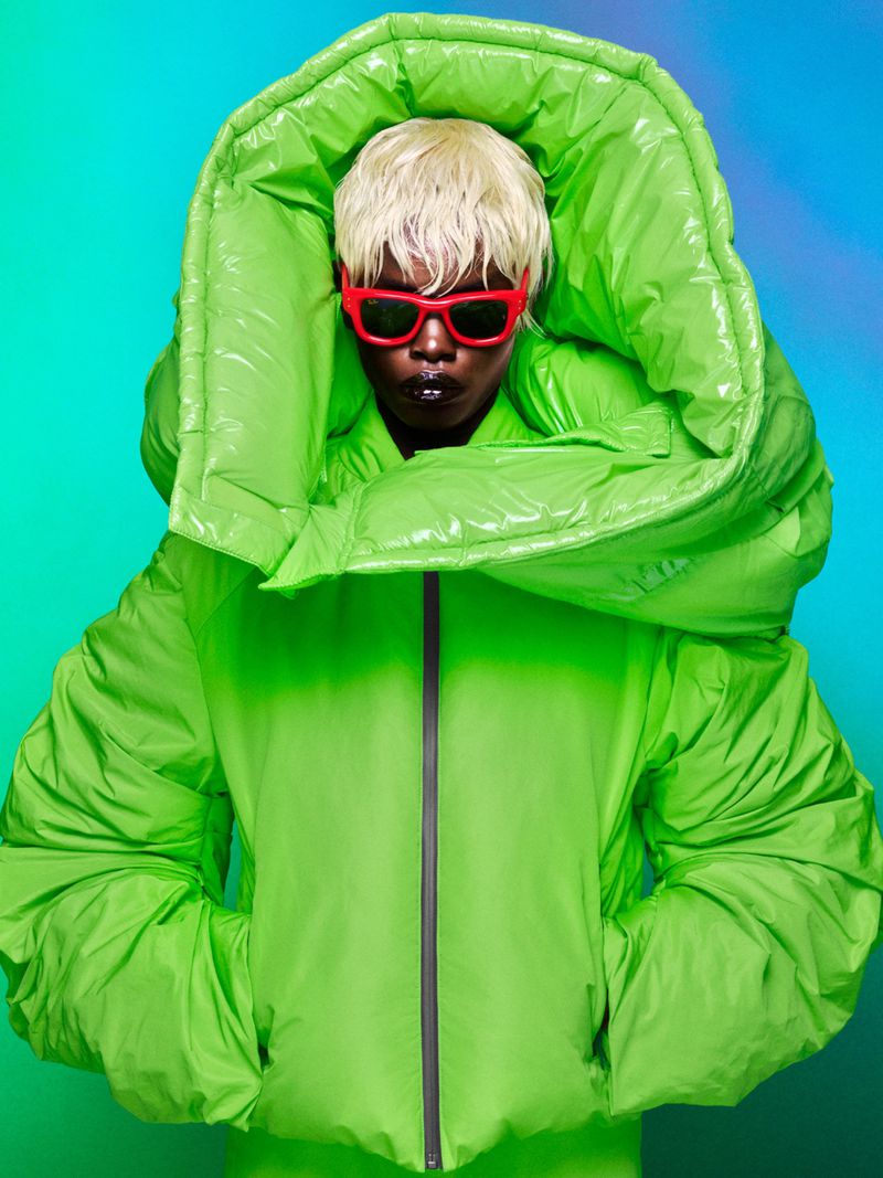 Person with platinum blonde hair, red glasses, and black lipstick wearing a large, glossy, bright green hooded puffer jacket in a studio setting.