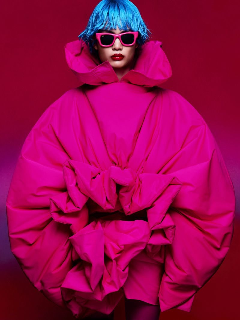 Person with blue hair wears hot pink dress and sunglasses against pink background in fashion style photograph. Color block.