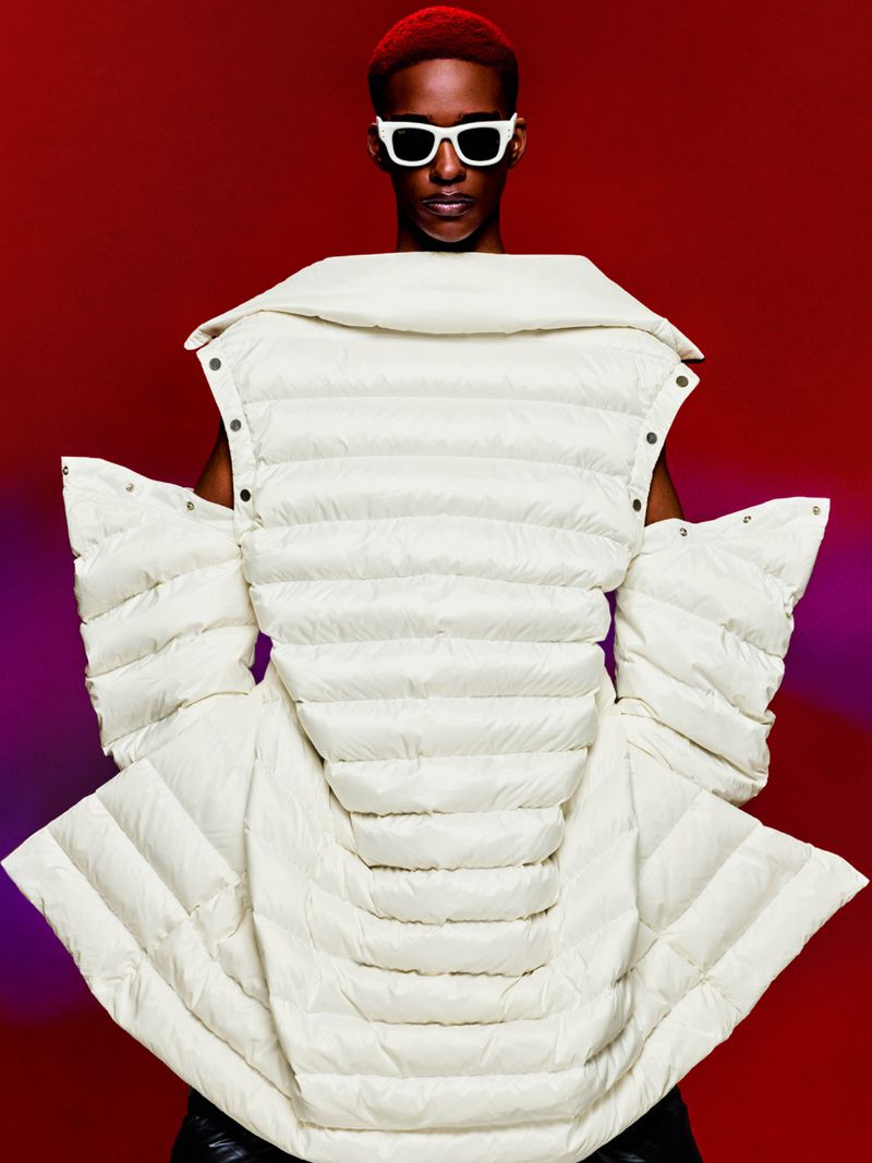 Man with reddish hair wearing white sunglasses and a cream-colored puffer jacket against a red and purple gradient background.