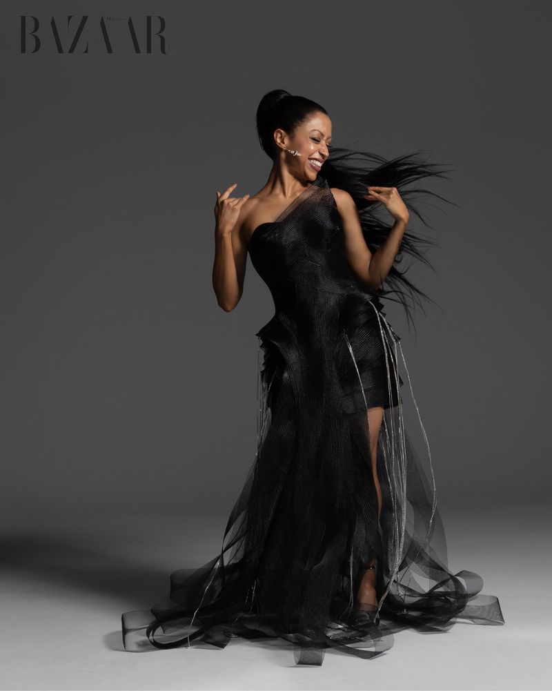 A smiling adult woman in a black, one-shoulder gown poses in a studio with flowing hair and upward-reaching arms against a gray backdrop.