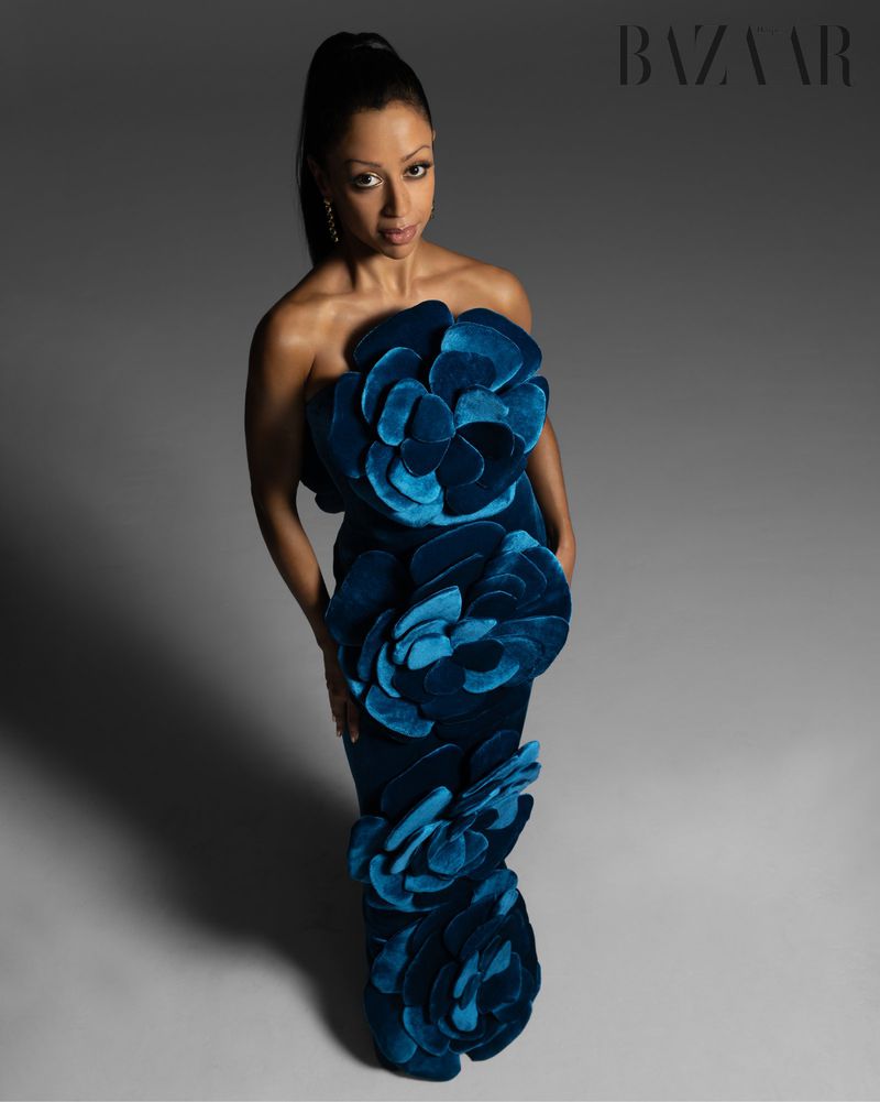 Elegant woman in a teal, strapless, flower-embellished gown posing in a photo studio with soft lighting against a gradient gray background.