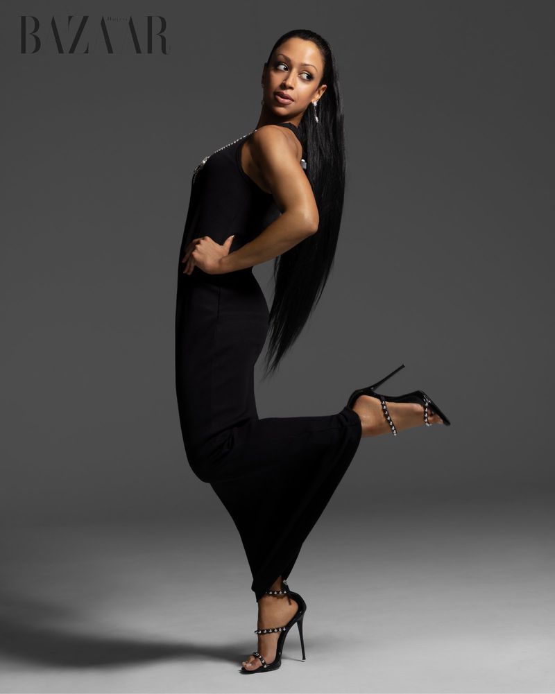 Liza Koshy posing in a long black dress and high heels against a neutral grey studio backdrop