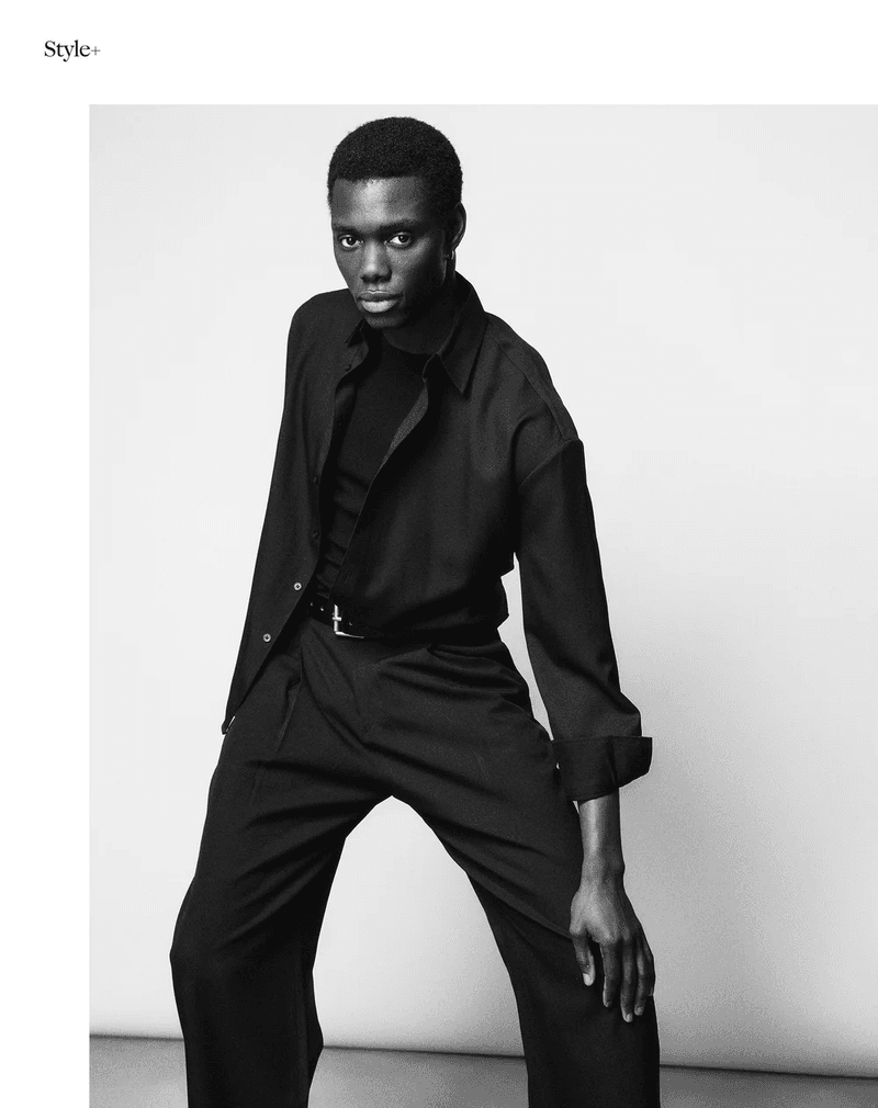 An adult Black male poses in a fashionable dark shirt and pants. The studio shot is simple and minimal against a neutral gradient backdrop.