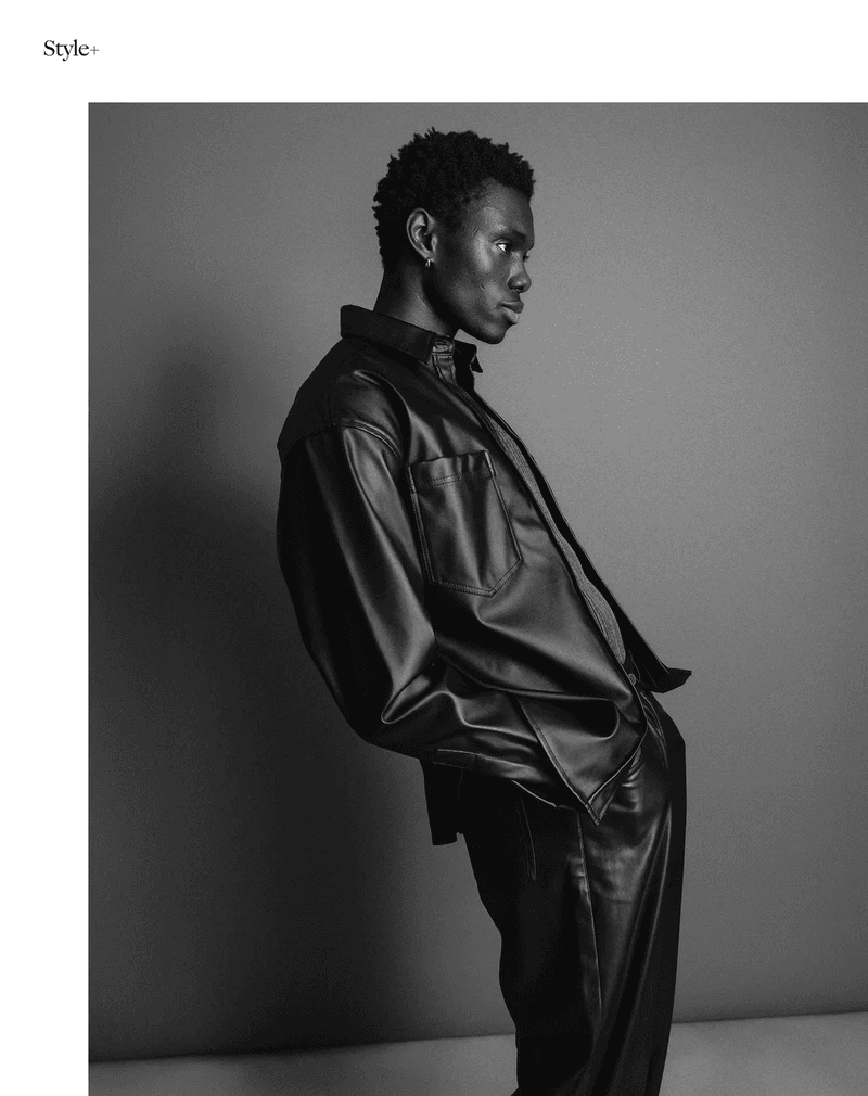 Monochrome portrait of a Black male model in a black leather-like shirt and pants, posed against a plain gray backdrop.