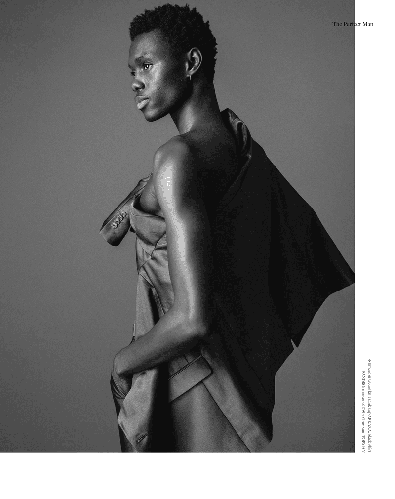 Monochrome fashion portrait of a Black male model with a jacket draped over his shoulder, posing against a grey background in a studio.