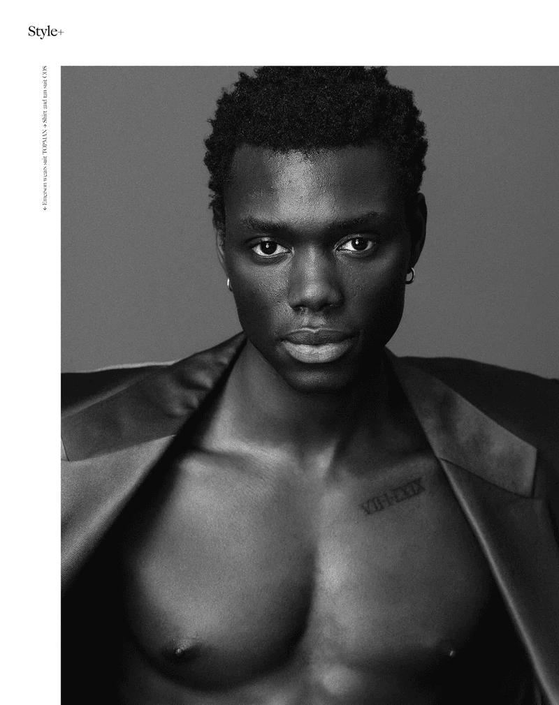 Striking black and white portrait of a muscular, shirtless Black man with a blazer, posing against a grey backdrop, highlighting his physique and style.