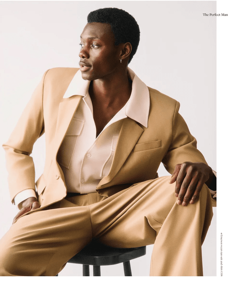 Portrait of a Black man in a tan suit sitting on a stool, looking off to the left, in a fashion editorial-style shot against a white background.