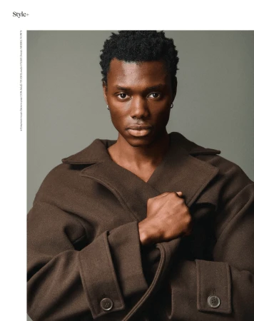 Portrait of a young adult man with dark hair wearing a brown wool coat against a neutral grey background in a studio setting.