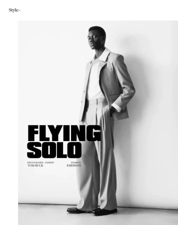 Black male model in light-colored suit poses against off-white wall in studio, showcasing editorial fashion and style.