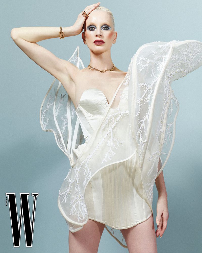 Woman in a white strapless dress with sculptural details, posing against a light blue background, gold jewelry, and magazine logo.
