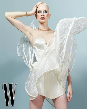 Woman in a white strapless dress with sculptural details, posing against a light blue background, gold jewelry, and magazine logo.