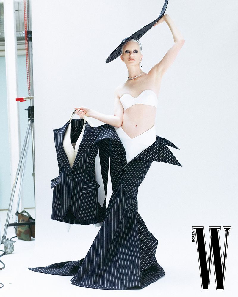 Fashion-forward studio shot of a woman in pinstripe skirt and white top, accessorized with a hat, holding a pinstripe jacket.