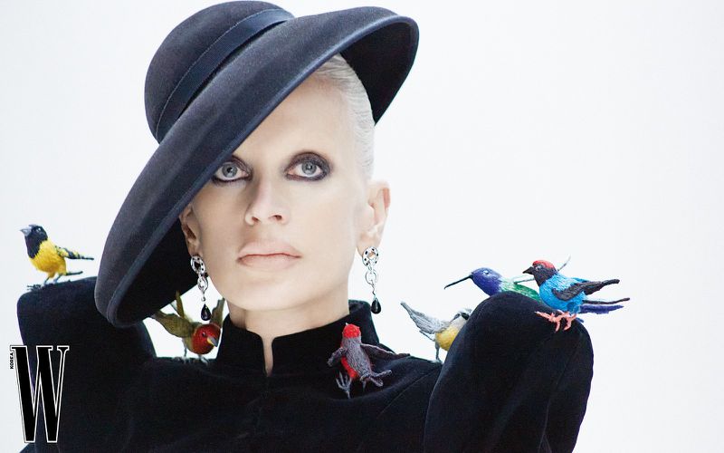 Fashion editorial shot of a woman in a black hat and jacket adorned with colorful bird brooches against a white background.