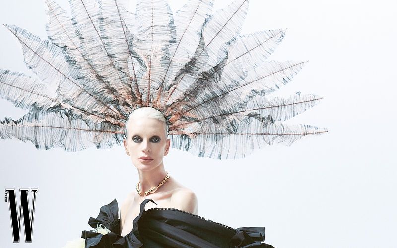 A fashion editorial shot of a fair-skinned adult female with platinum hair, wearing a feather headpiece, black top, and gold necklace.