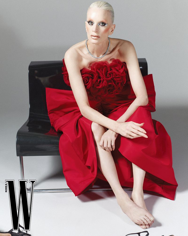 Fashion editorial shot of a fair-skinned adult woman in a red strapless gown sitting on a black chair against a grey backdrop.