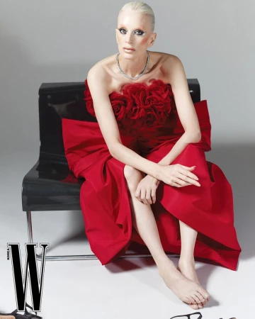Fashion editorial shot of a fair-skinned adult woman in a red strapless gown sitting on a black chair against a grey backdrop.