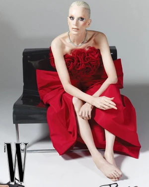 Model Kristen McMenamy wearing a red strapless dress seated on a black chair against a grey seamless background