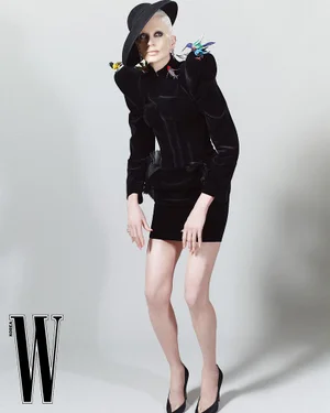 Model Kristen McMenamy in a structured black velvet mini dress, black hat, and bird brooches on a grey studio backdrop.