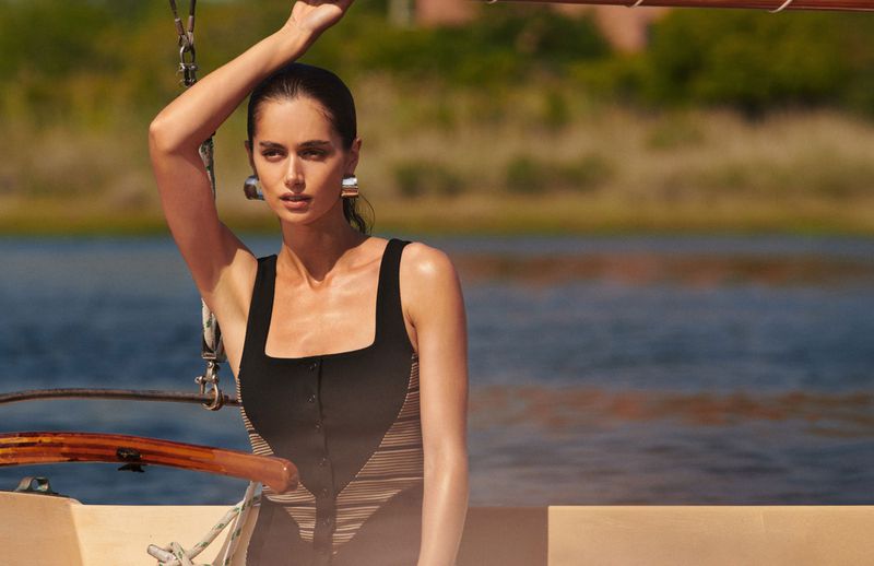 Woman in a bathing suit poses on a sailboat with a distant shoreline and blue water in the background in an outdoor fashion shot.