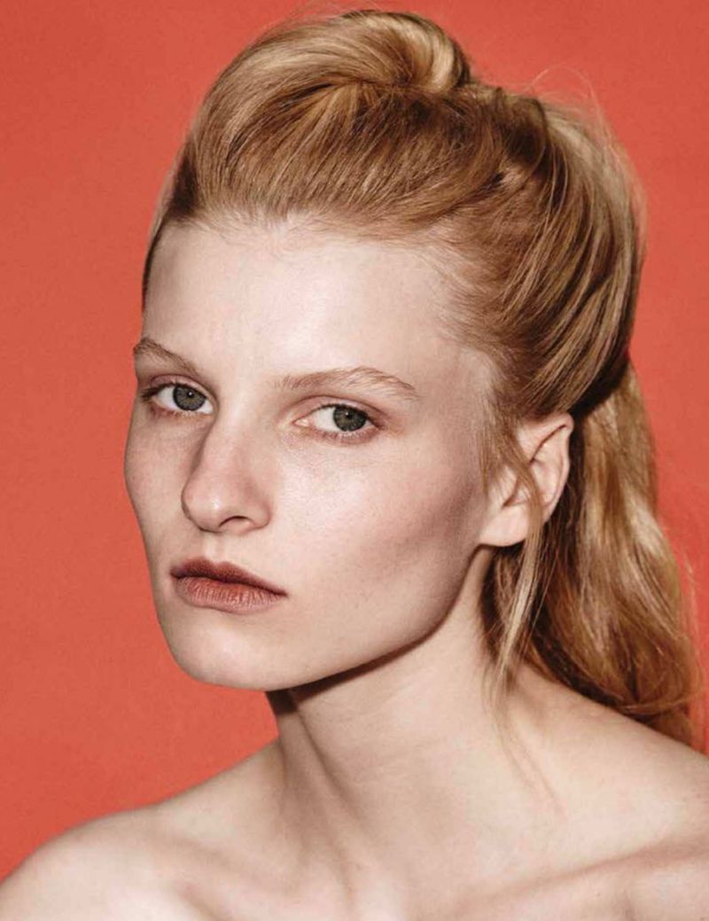 Portrait of an adult woman with light blonde hair against a plain coral backdrop in a beauty or fashion context, close-up studio shot.
