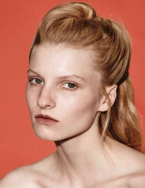 Portrait of an adult woman with light blonde hair against a plain coral backdrop in a beauty or fashion context, close-up studio shot.