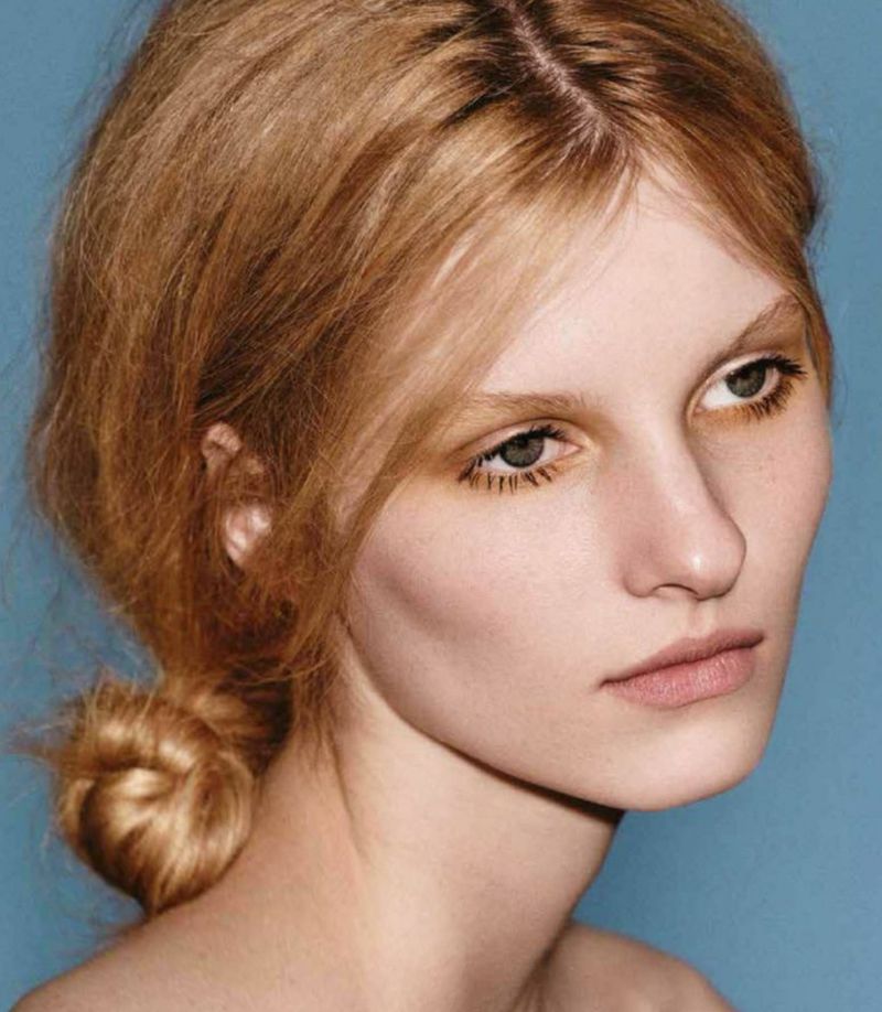 Close-up portrait of a blond adult woman with makeup against a blue background in a beauty or fashion editorial. Simple hair styling.