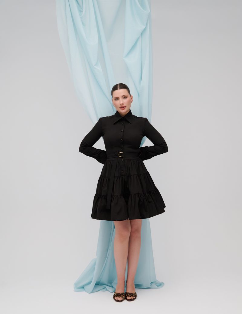 Full-length portrait of a woman in a black dress with a belt, standing against a light blue draped fabric and a white background.